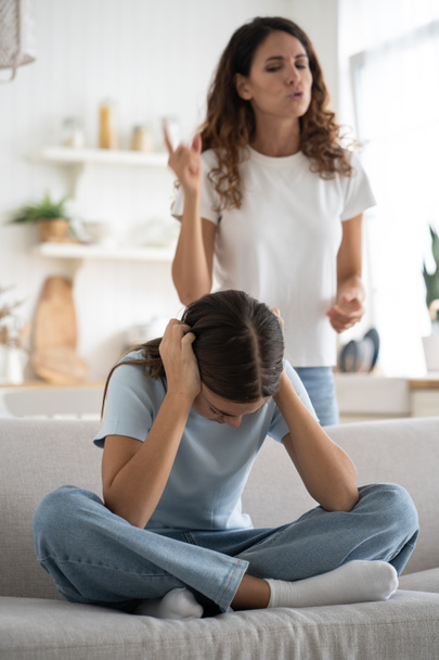 Sad teenage girl sits cross-legged on couch and covers ears with hands not wanting to listen to mother reproaches. Nervous woman looking at daughter scolding after teacher call from school. Parenting. - Photo, Image