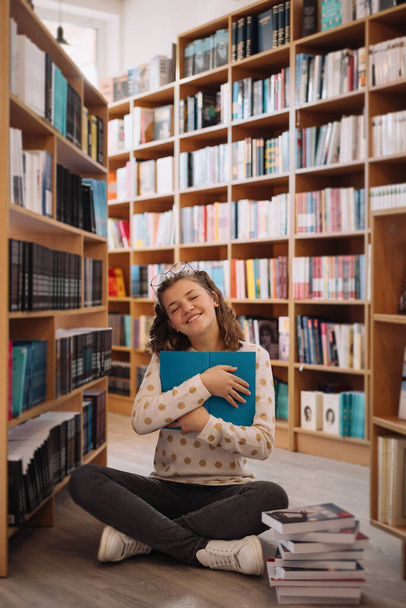 本の山の中で10代の女の子。若い女の子が背景に棚のある本を読んでいる。彼女は本の山に囲まれている。予約日. - 写真・画像