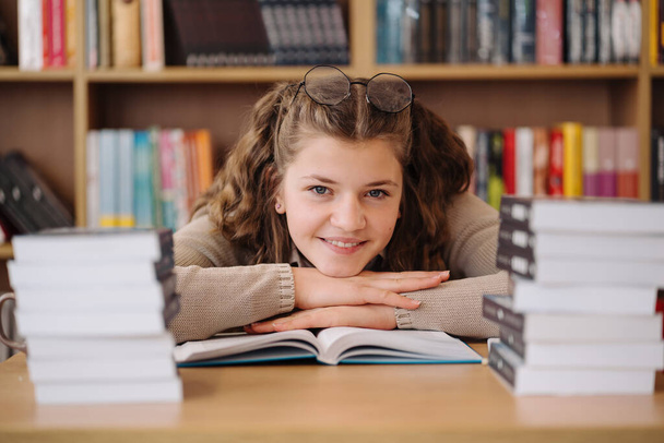 Ελκυστική ευτυχισμένη νεαρή μαθήτρια που σπουδάζει στη βιβλιοθήκη του κολεγίου, κάθεται στο γραφείο - Φωτογραφία, εικόνα