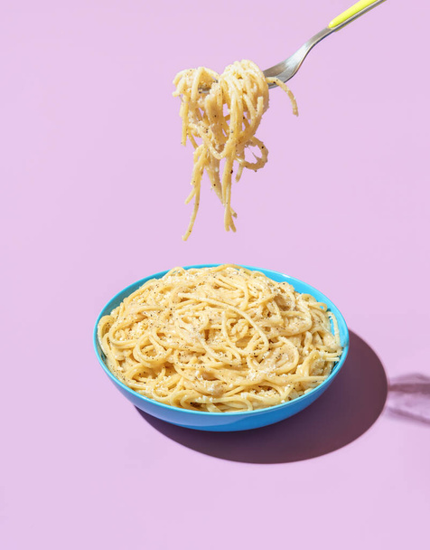 Pasta Cacio e Pepe in a blue bowl isolated on a purple background. Taking spaghetti with a fork from a bowl in bright light against a colorful background. - Photo, Image