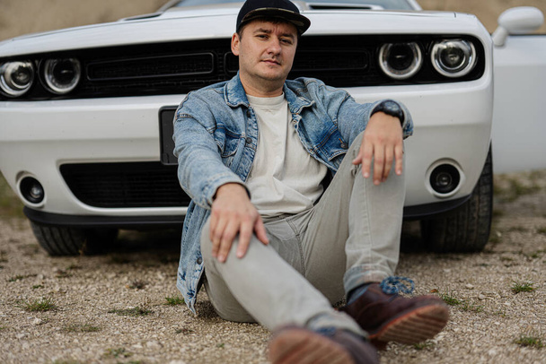 Handsome man in jeans jacket and cap sitting near his white muscle car in career. - Foto, Bild
