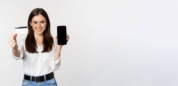 Portrait of female model showing credit card with smartphone screen, recommending application, standing over white background. - Φωτογραφία, εικόνα