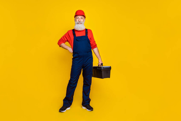 Full size photo of positive professional builder long beard red headgear hold tool box hand on waist isolated on yellow color background. - Foto, Imagem