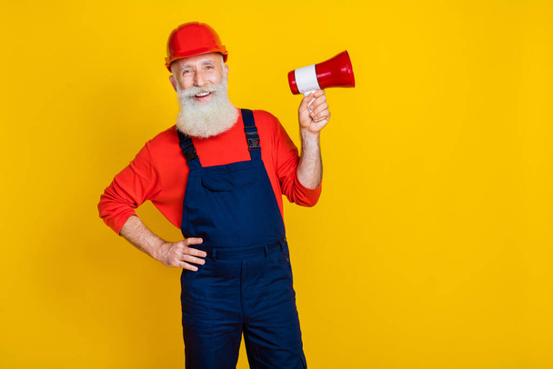 Photo of happy smiling age man wear overall uniform red hard hat holding bullhorn empty space isolated yellow color background. - Φωτογραφία, εικόνα