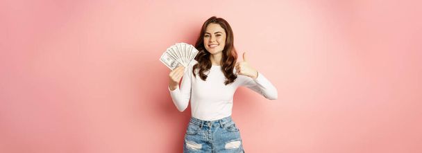 Jednoduché peníze a půjčka. Usmívající se krásná žena ukazující peníze se spokojeným obličejem, ukazující palce nahoru, stojící na růžovém pozadí. - Fotografie, Obrázek