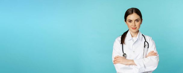 Medical staff and doctors concept. Young smiling female doctor, healthcare worker in white coat and stethoscope, looking confident, waiting patients, blue background. - Foto, afbeelding