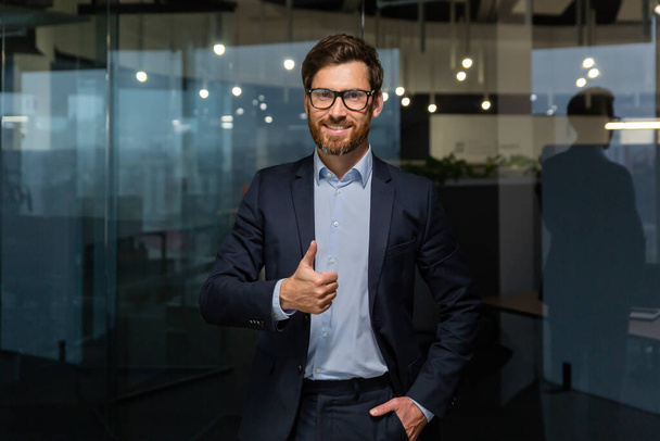 Portrait of successful mature businessman boss, manager in business suit glasses and beard looking at camera and smiling standing near window, showing thumbs up, sign of success and achieving goals. - 写真・画像