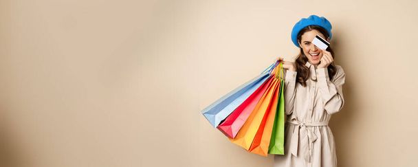 Portrat of trendy feminine girl posing with shopping bags from store and credit card, paying contactless, buying with discount on sale, beige background. - Foto, Imagem
