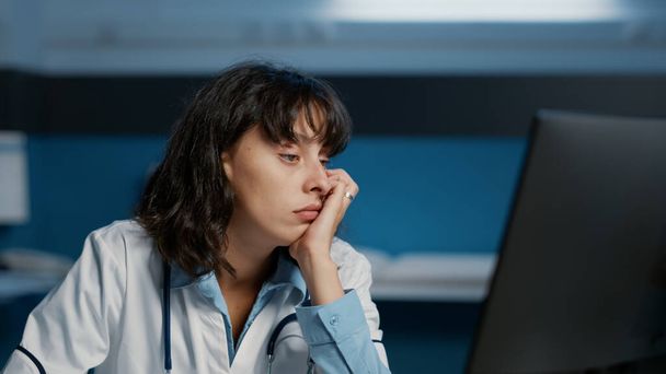 Tired doctor woman falling asleep while working late at night in hospital office, analyzing patient disease report on computer typing medical expertise. Stressed medic planning health care treatment - 写真・画像