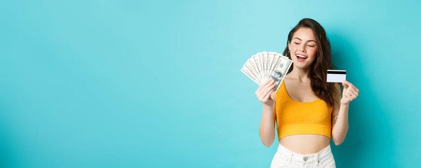 Όμορφο χαμογελαστό κορίτσι που κρατά πιστωτική κάρτα, που φαίνεται ευχαριστημένη και αποφασισμένη σε δολάρια χαρτονομίσματα, στέκεται απέναντι σε μπλε φόντο. - Φωτογραφία, εικόνα