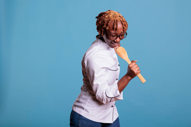 Cheerful woman holds a spatula in hand simulating microphone, dances and sings with excitement. Energetic female chef wears white work jacket in studio shot against blue background. - Photo, Image