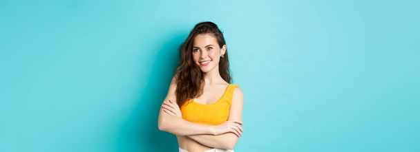 Sommer- und Lifestyle-Konzept. Stilvolle moderne Frau in abgeschnittenem Top, selbstbewusst wirkend, verschränkte Arme auf der Brust und entzückt lächelnd, vor blauem Hintergrund stehend. - Foto, Bild
