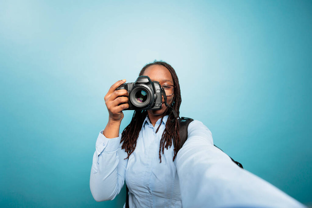 Νεαρή ενήλικη Αφροαμερικανή γυναίκα που δείχνει τη συσκευή DSLR στην κάμερα ενώ τραβάει μια φωτογραφία σε μπλε φόντο. Δημιουργικός ερασιτέχνης φωτογράφος φωτογραφίζει με σύγχρονη φωτογραφική μηχανή για τα μέσα κοινωνικής δικτύωσης. - Φωτογραφία, εικόνα