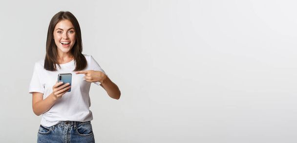Excited and pleased brunette girl pointing finger at smartphone screen. - Foto, imagen