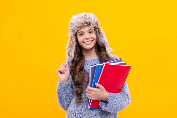 happy teen girl wear earflap hat with notebook on yellow background, education. - Fotó, kép
