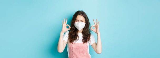 Covid, koncepcja zdrowia i pandemii. Zadowolona piękna dziewczyna w respiratorze, maska medyczna wykazująca zgodę na znak, uisng środki zapobiegające koronawirusowi, niebieskie tło. - Zdjęcie, obraz