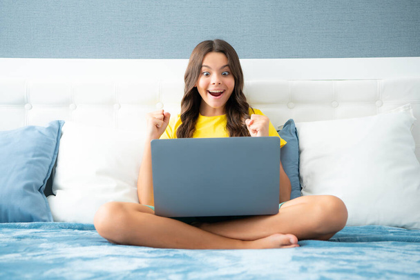 Tiener schoolmeisje studeert thuis op bed met laptop. Expressieve emotionele opgewonden tiener meisje - Foto, afbeelding