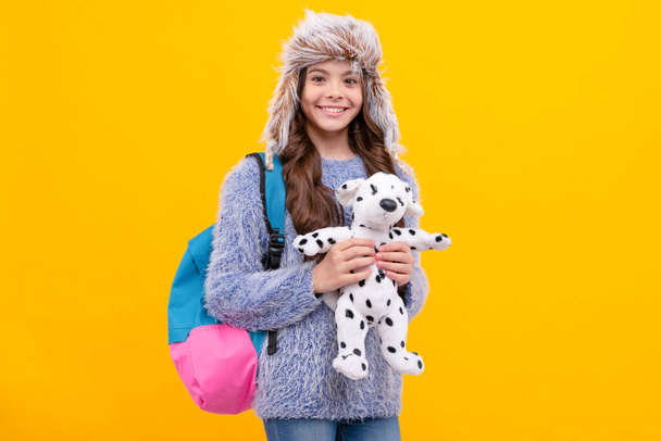 child wearing warm clothes carry backpack. express positive emotion. winter fashion. happy kid with curly hair in hat hold toy. back to school. teen girl in knitwear on yellow background. - Photo, Image