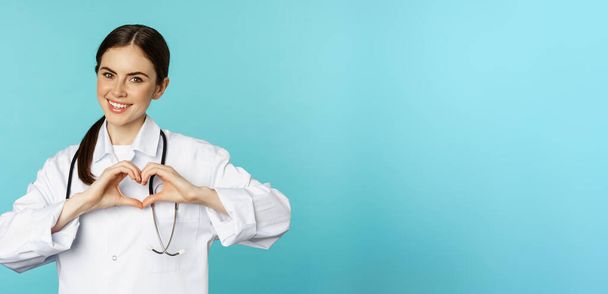 Nuori lääkäri, nainen lääkäri, joka osoittaa sydäntä, rakkauden merkki ja hymyilevä, hoitaa potilaita klinikalla, seisoo valkoisessa takissa sinisellä taustalla. - Valokuva, kuva