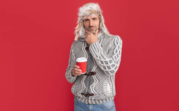 Σκέφτομαι τον Χάρρυ να πίνει καφέ. ανδρική μόδα μοντέλο κρατήσει κύπελλο. χειμερινή μόδα. Ο πρωινός καφές παίρνει μακριά το κύπελλο. Γκρινιάρης τύπος με πουλόβερ και καπέλο σε κόκκινο φόντο. - Φωτογραφία, εικόνα
