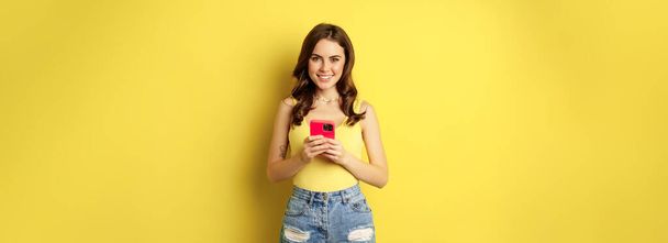 Κυτταρική τεχνολογία και online αγορές έννοια. Ενθουσιαστικό όμορφο κορίτσι στέκεται με smartphone και χαμογελαστό, στέκεται απέναντι σε κίτρινο φόντο. - Φωτογραφία, εικόνα