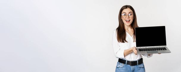 Молода жінка в окулярах демонструє екран ноутбука, демонструючи рекламу на комп'ютері, веб-сайті або магазині, стоячи на білому тлі
. - Фото, зображення
