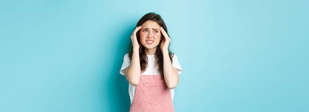 Femme ayant une migraine douloureuse, touchant la tête et fronçant les sourcils, regardant le coin supérieur gauche, souffrant de maux de tête, debout sur fond bleu. - Photo, image