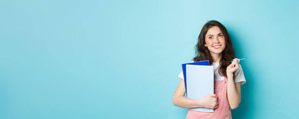 Gyönyörű lány diák néz figyelmes a felső másolás helyet, álló notebook házi feladatot és mosolygós töprengő, álló ellen kék háttér. - Fotó, kép