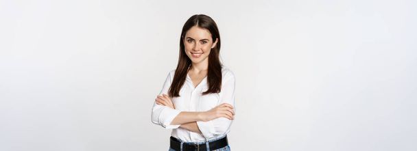 Uśmiechnięta młoda bizneswoman, przedsiębiorczyni w białej koszuli, krzyżujące ramiona na klatce piersiowej jak profesjonalistka, stojąca nad studiem. - Zdjęcie, obraz