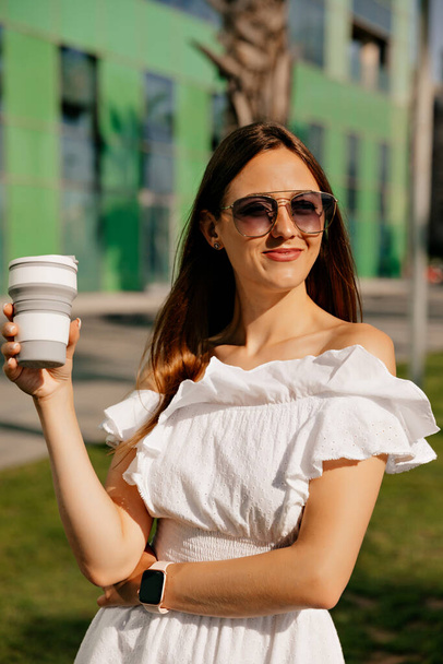 Close up retrato de jovem mulher elegante com cabelos longos soltos vestindo vestido de verão branco e óculos de sol olhando para o lado com café pela manhã.  - Foto, Imagem