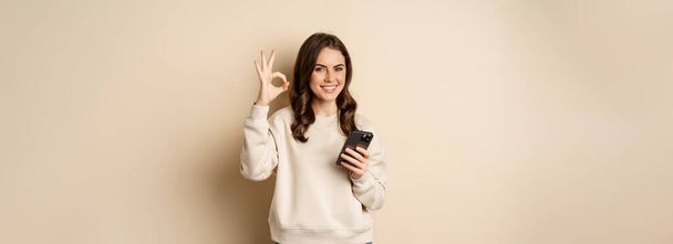 Beautiful woman holding cellphone, mobile phone and okay sign, recommending application, shopping app, standing over beige background. - Fotó, kép