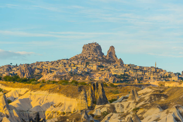 Buildings carved into rocky mountain in Cappadocia, Turkey. Rocky hills and grasslands with clear blue sky and bright yellow summer morning sunlight. Tourist destination. Horizontal shot. High quality - Photo, Image