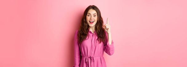 Excited beautiful girl with make up on, having an idea, raising finger up in eureka sign and smiling, saying her plan, standing against pink background. - Фото, изображение