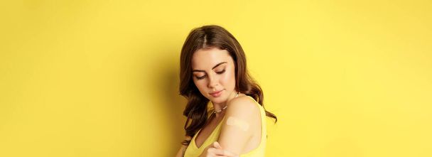 Vacunación con Covid-19. Retrato de cerca de una joven milenaria que mira un parche en el hombro después de la vacuna de la inyección del coronavirus, de pie sobre un fondo amarillo. - Foto, imagen