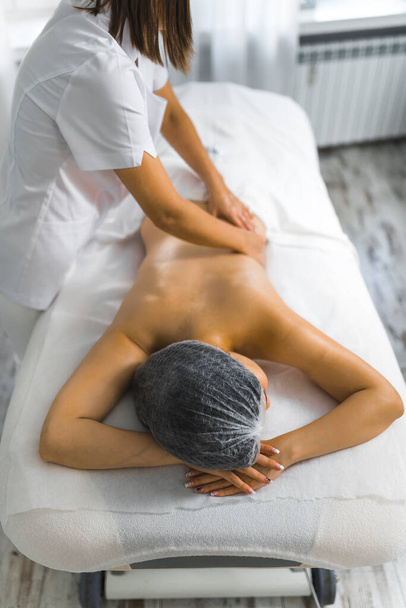 Vertical indoor shot of skinny young Asian woman lying on her stomach on massage bed being massaged by unrecognizable caucasian female massage therapist. SPA day. High quality photo - Фото, изображение