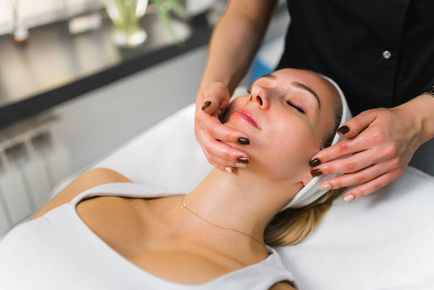 Unrecognizable caucasian aesthetician performing gentle face massage on her beautiful young female client. Indoor closeup shot. . High quality photo - Photo, Image