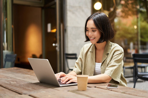 Jonge Aziatische vrouw, digitale nomade die op afstand werkt vanuit een café, koffie drinkt en laptop gebruikt, glimlacht. - Foto, afbeelding