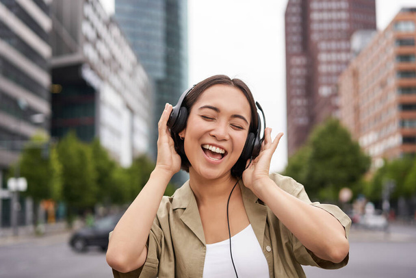 Dancing girl feeling happy in city. Asian woman dancing and listening music in headphones, posing on street. - Photo, Image