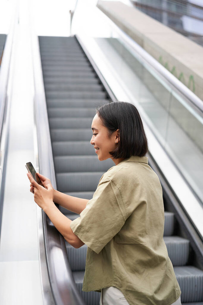 Mladá Asiatka jde na eskalátor, drží smartphone, usmívá se, když jde po městě. Kopírovat prostor - Fotografie, Obrázek