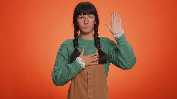 Vannon olevani rehellinen. Vilpitön vastuullinen nuori nörtti nainen nostaa kätensä vannomaan valan, lupaamalla olla rehellinen ja kertoa totuuden, pitää käsi rinnassa. Latina tyttö eristetty studio oranssi tausta - Valokuva, kuva