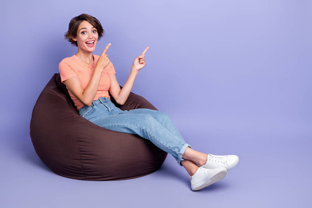 Full body photo of charming young girl sit comfy beanbag point empty space wear trendy striped garment isolated on violet color background. - Φωτογραφία, εικόνα