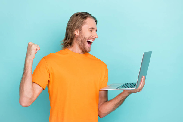 Photo of positive lucky man wear orange t-shirt reading modern gadget rising fist empty space isolated turquoise color background. - Zdjęcie, obraz