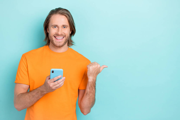Photo of pretty cute guy dressed orange t-shirt looking pointing thumb empty space typing modern device isolated teal color background. - Foto, Bild