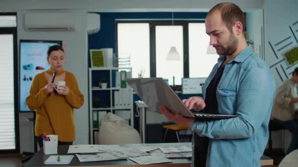 Portrait of casual business man using laptop and smiling at camera while coworker holding cup looks at papers with charts. Confident startup employee working relaxed in busy office of small company. - Záběry, video