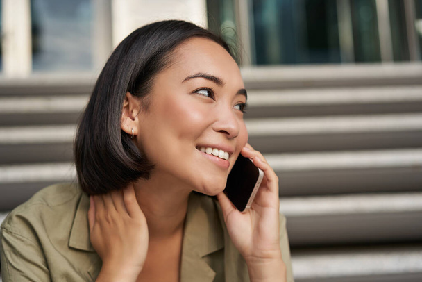 Tecnología celular. Sonriente chica asiática habla en el teléfono móvil, hace una llamada. Mujer joven con teléfono se sienta en las escaleras. - Foto, imagen