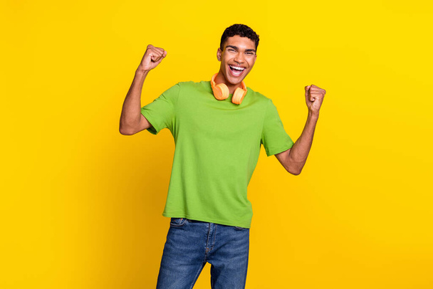 Photo portrait of handsome young man raise fists scream hooray earphones wear stylish green clothes isolated on yellow color background. - Foto, afbeelding