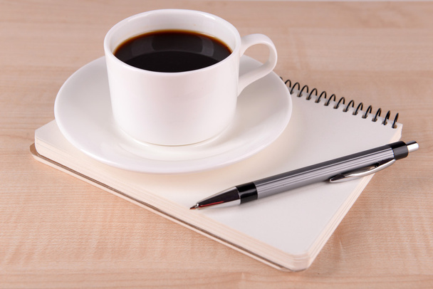 Cup of coffee on saucer with notebook and pen on wooden table background - Фото, зображення