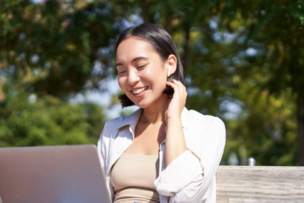 Gyönyörű fiatal koreai nő vezeték nélküli fülhallgató, video chat, munka a szabadban a parkban laptop, mosolygós és boldognak tűnik. - Fotó, kép