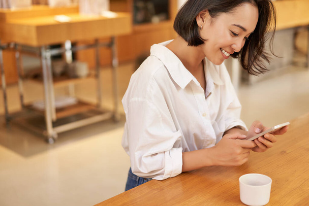 Retrato de mujer asiática joven, sentada en la cafetería, sosteniendo el teléfono inteligente, charlando y mensajería mientras bebe café. - Foto, Imagen
