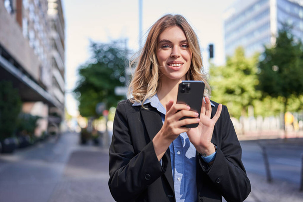 Retrato de una mujer de negocios sonriente usando teléfono móvil mientras está de pie al aire libre cerca de edificios de oficinas. - Foto, Imagen